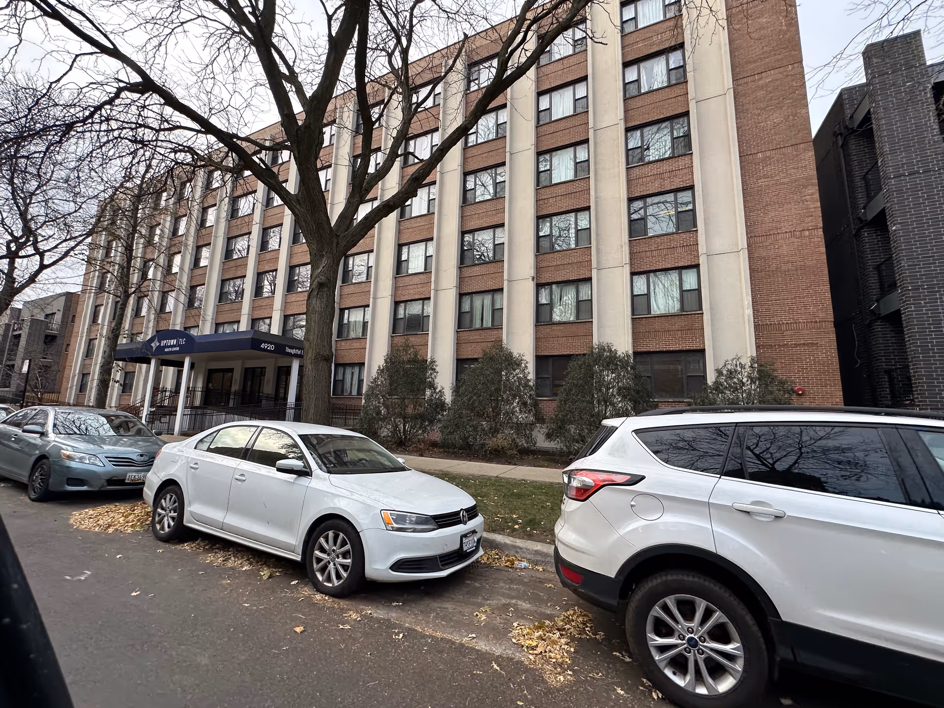 Front exterior of a multi-story brick care facility with a canopy entrance, parked cars, and a large tree in front.