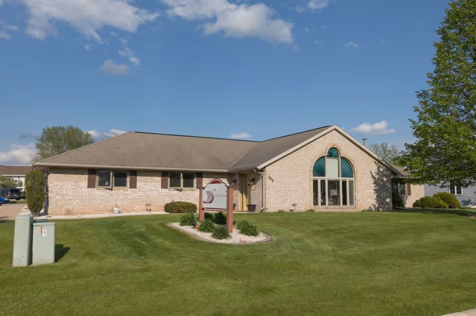 Single-story brick assisted-living building with large arched front windows, a manicured lawn, and a wooden sign near the entrance.