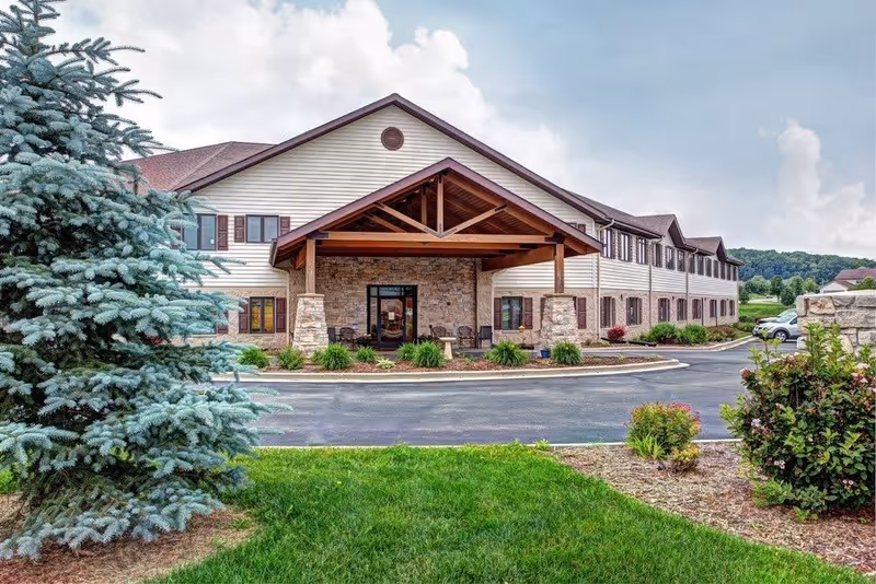Exterior view of a two-story senior living facility building with a covered entrance supported by stone pillars. The building has beige siding with brown shutters and a stone facade around the entrance. There is a paved driveway and landscaped greenery including a large evergreen tree and bushes in the foreground under a partly cloudy sky.