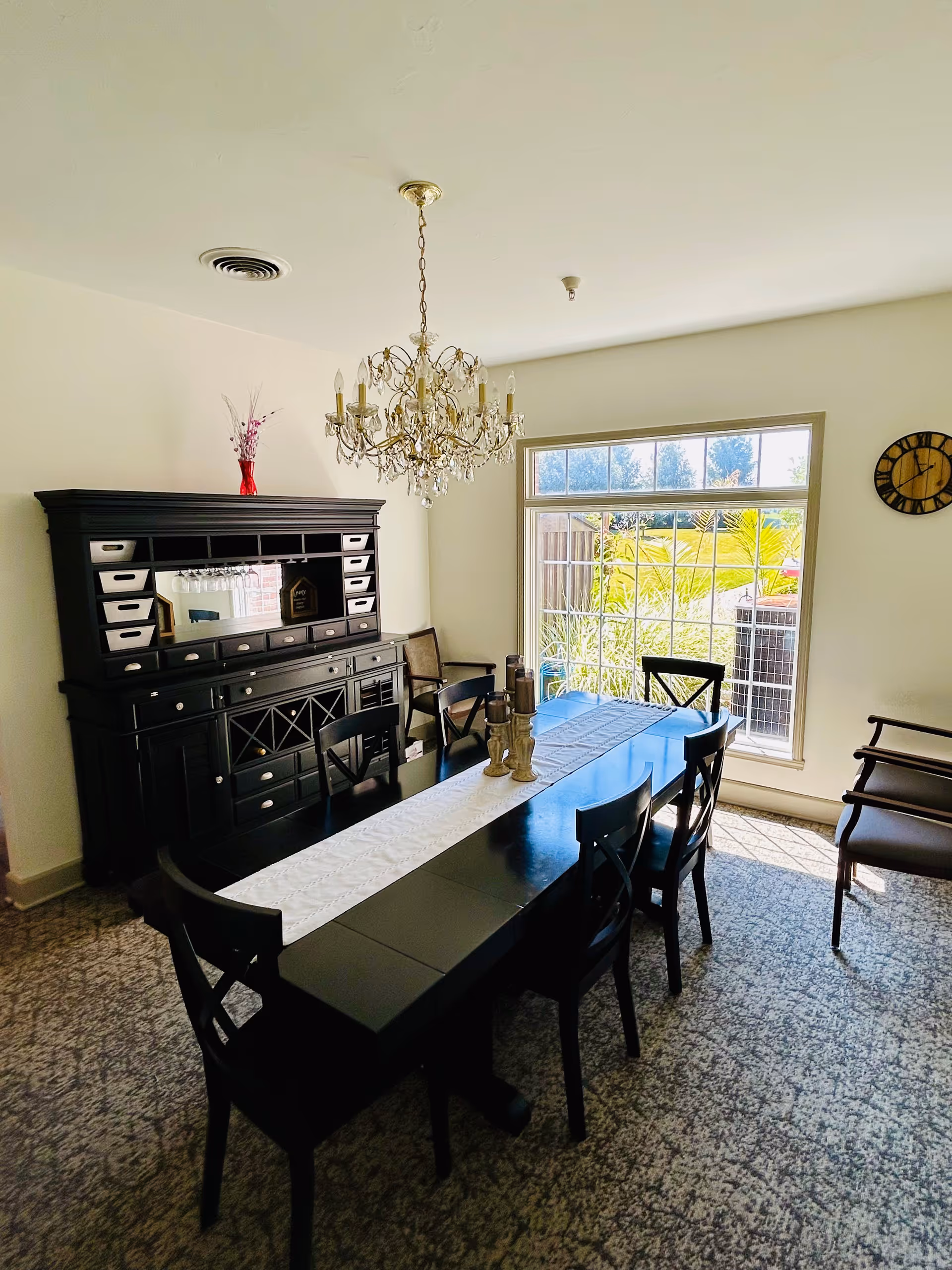 Bright dining room with a long black table and chairs, a crystal chandelier, a dark hutch, and a large window overlooking greenery.