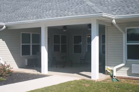 Covered outdoor patio area attached to a building with several windows and chairs, surrounded by a small lawn and a concrete walkway.