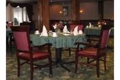 A dining room with tables covered in green tablecloths, set with white napkins, cups, and plates. The chairs have wooden frames with red cushioned seats and backs. The room has warm lighting and a carpeted floor with a patterned design.