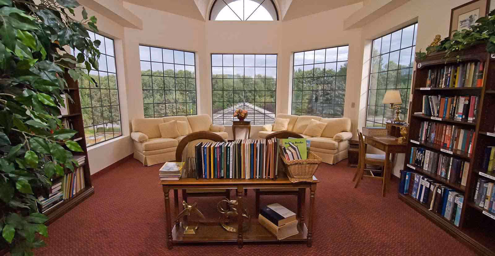 A cozy senior living library room with large windows showing a green outdoor view. The room features two beige sofas with cushions, a wooden table with books arranged on it, bookshelves filled with books, a small desk with a lamp, and a potted plant on the left side.
