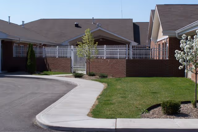 Exterior view of a brick senior living building with a curved driveway, sidewalk, lawn, and gated fenced entrance.