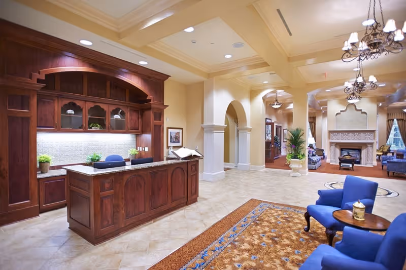 Reception lobby with a wooden front desk, blue upholstered chairs, chandeliers, and a fireplace in the background.