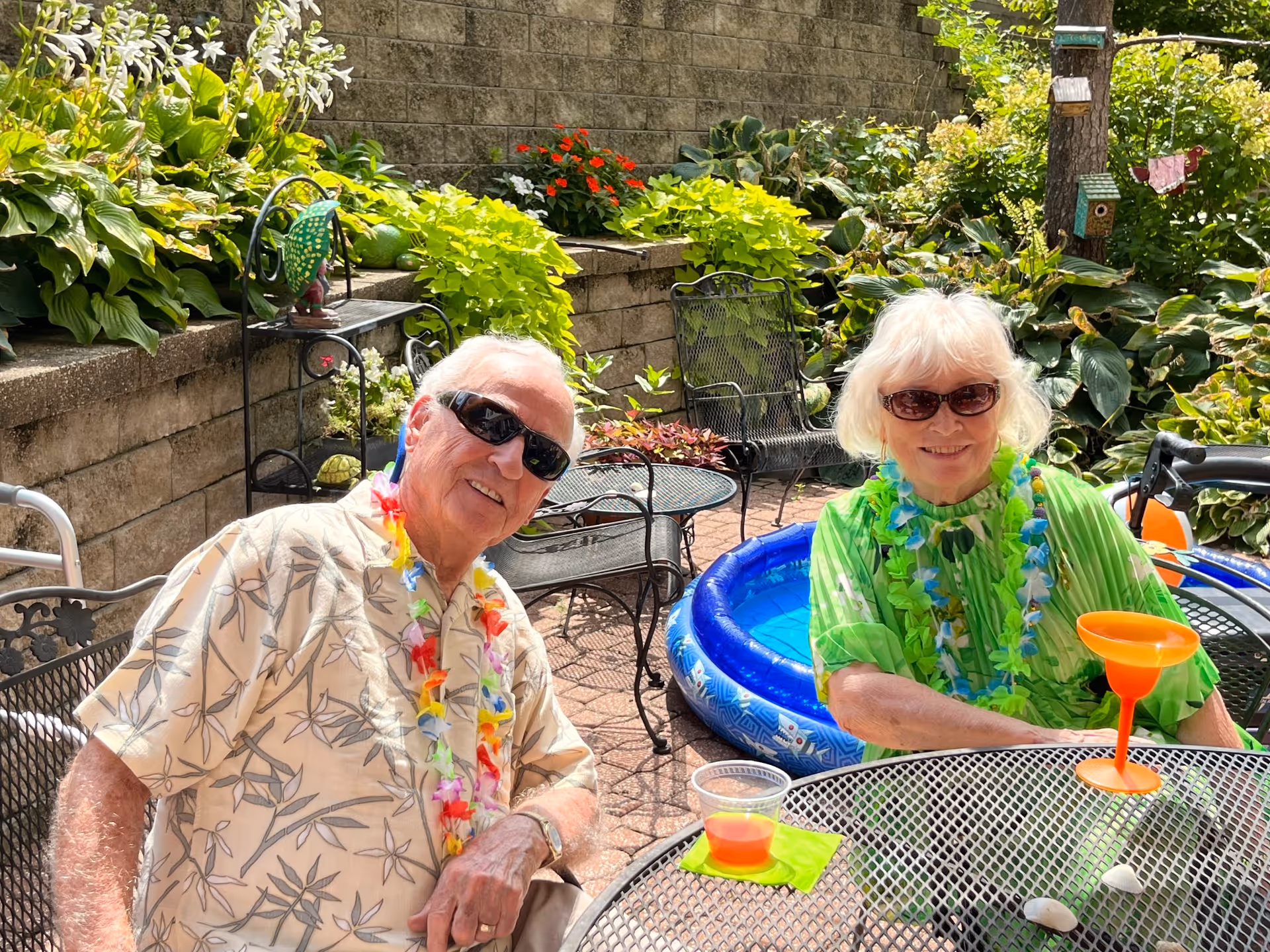 Two elderly people wearing sunglasses and colorful leis sitting at a metal outdoor table with drinks. Behind them is a small inflatable pool and lush green plants along a stone wall.