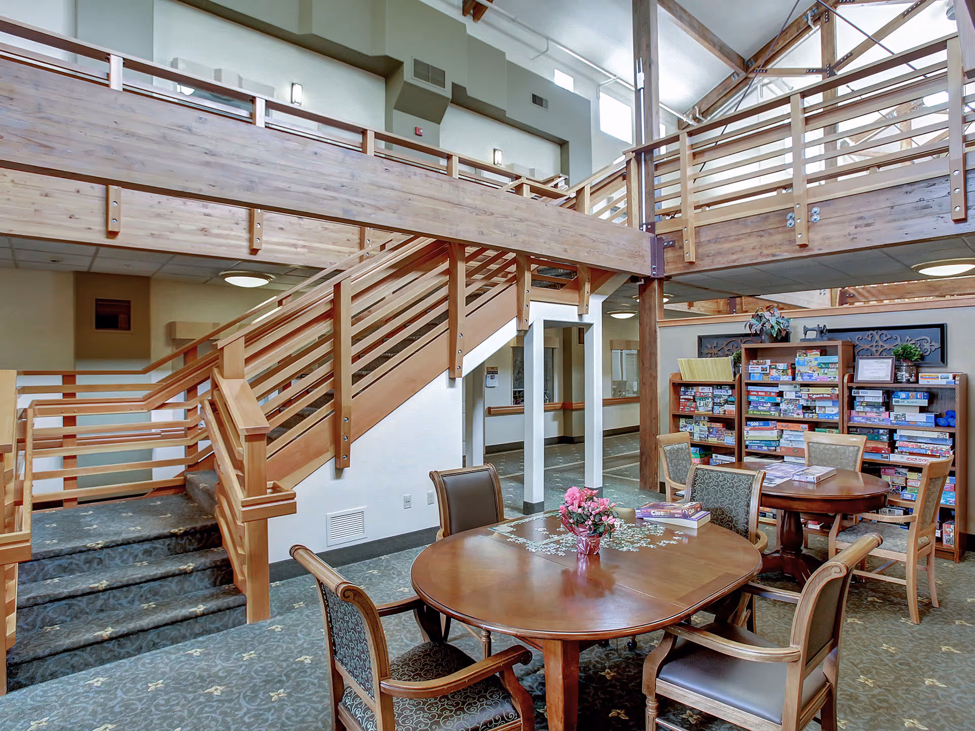Interior of a senior living facility with a wooden staircase and railing, a carpeted floor, and two round wooden tables surrounded by chairs. One table has a vase with pink flowers and a puzzle in progress, while the other table has board games on it. Behind the tables is a large bookshelf filled with various board games and puzzles. The space has high ceilings with exposed beams and windows near the ceiling allowing natural light.