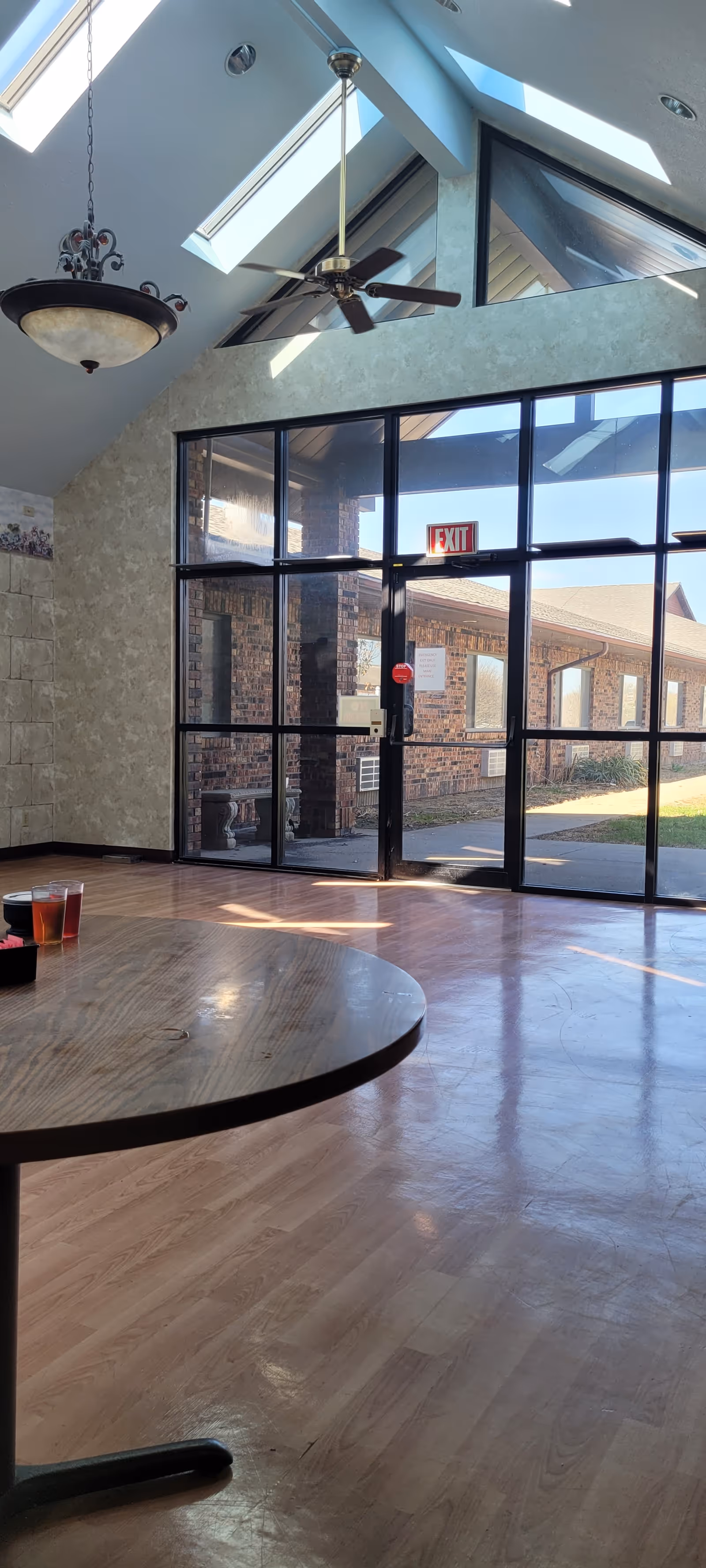 Sunlit common room with a vaulted skylighted ceiling, a ceiling fan, a round wooden table in the foreground and large glass doors leading outside.
