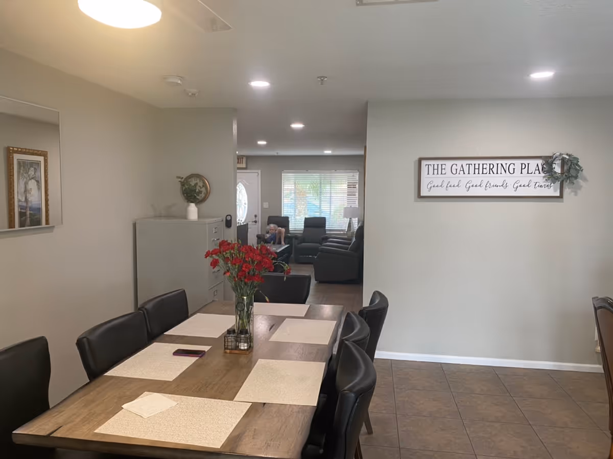 Interior view of a dining area with a wooden table set with placemats and a vase of red flowers in the center. Black chairs surround the table. In the background, there is a living room area with recliner chairs and a window with blinds. A sign on the wall reads 'THE GATHERING PLACE Good food, Good friends, Good times.'