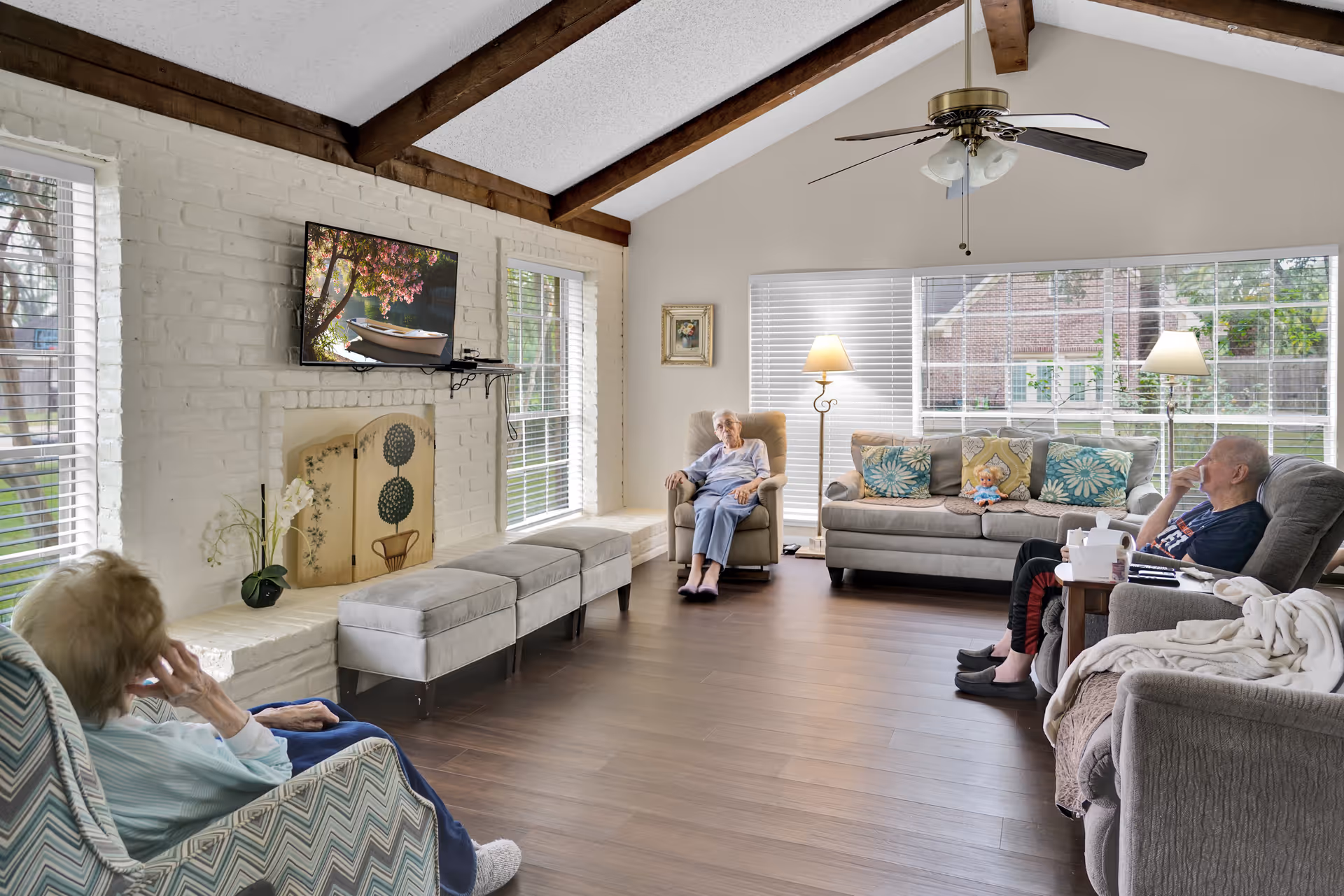 Bright living room in a memory-care facility with three seated residents, sofas and armchairs, a wall-mounted TV above a white brick fireplace, large windows, and exposed wooden ceiling beams.