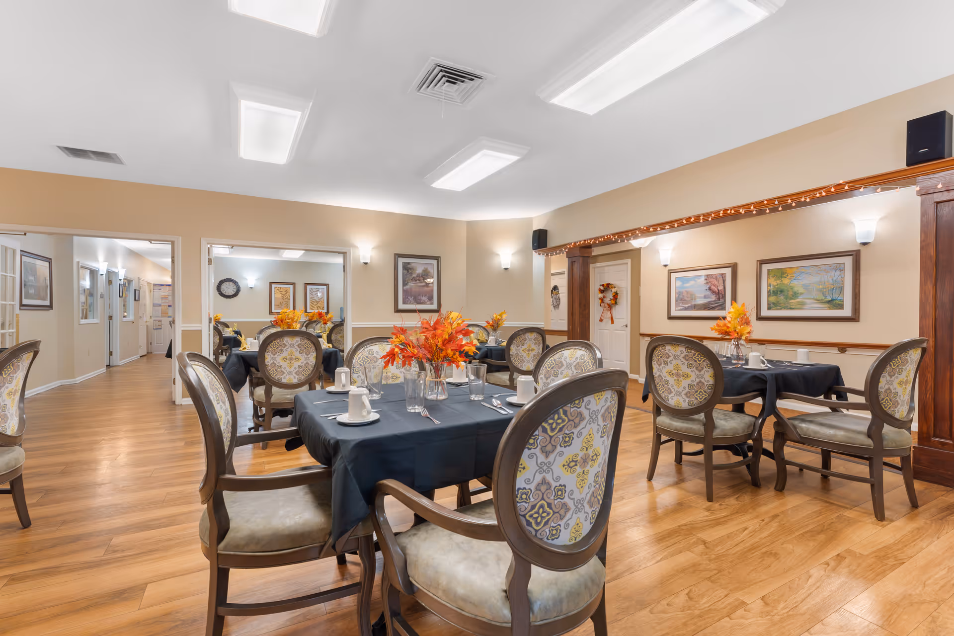 A dining room in a senior living facility with multiple tables covered in dark tablecloths, each set with cups, plates, and silverware. The chairs have patterned upholstery with a floral design. The room has wooden flooring, beige walls adorned with framed paintings, and soft lighting from wall sconces and ceiling lights. There are autumn-themed floral centerpieces on the tables and string lights along a wooden beam.