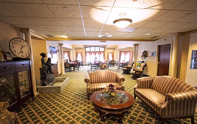 Spacious senior living common room with striped sofas and a coffee table in the foreground and dining tables near windows in the background.