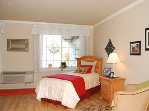 Sunlit single bedroom with a bed dressed in white and red linens, wooden nightstand with lamp, window and a chair.
