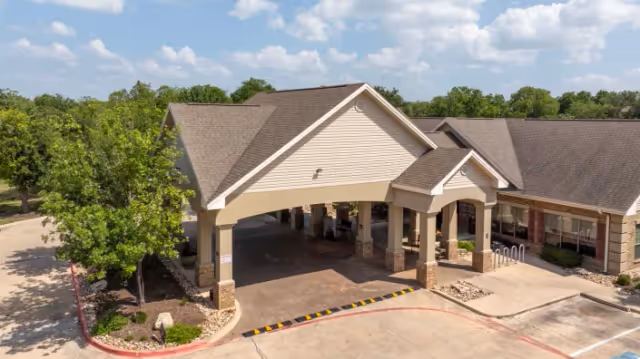 Aerial view of a senior living facility's front entrance featuring a covered porte-cochère, beige siding, stone columns, and a drive-up drop-off area.