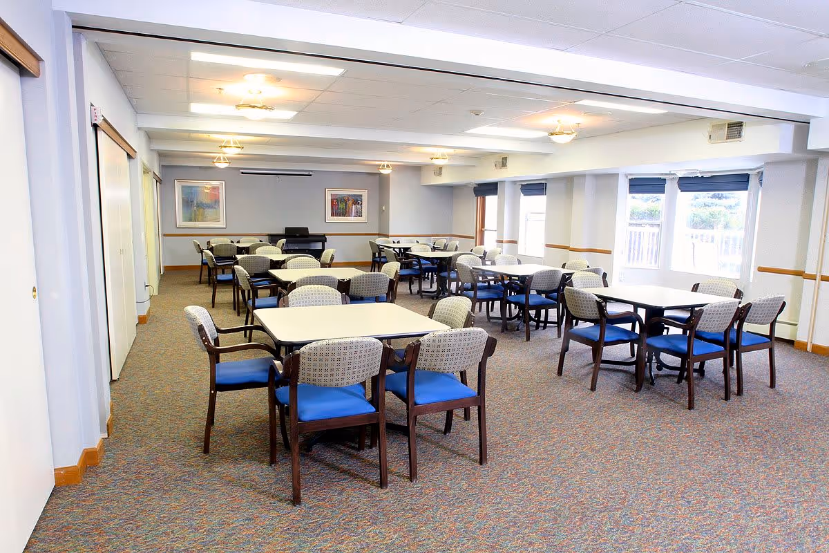 A bright and spacious dining room in a senior living facility with multiple square tables surrounded by chairs with blue cushions and patterned backs. The room has carpeted floors, large windows with blue blinds, framed artwork on the walls, and ceiling lights providing illumination.
