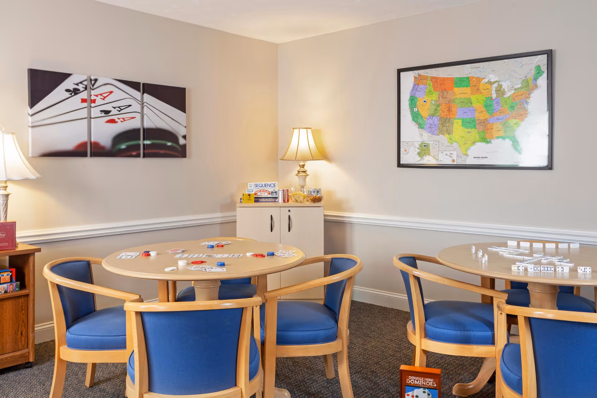A cozy game room with two round wooden tables surrounded by blue cushioned chairs. One table has playing cards and chips, while the other has dominoes set up. The walls are beige with white trim, decorated with a large map of the United States and a three-panel artwork of playing cards. A lamp on a small cabinet provides warm lighting.