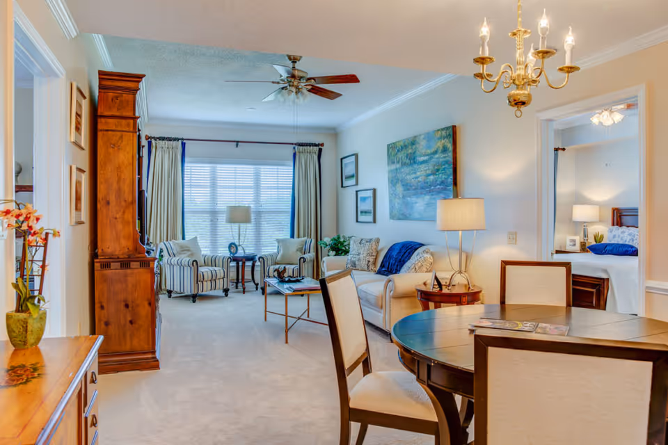 A bright and cozy living and dining area in a senior living facility. The living room features two striped armchairs, a white sofa with cushions, a coffee table, and a large window with curtains letting in natural light. The dining area has a round wooden table with four upholstered chairs. A ceiling fan and a chandelier provide lighting, and artwork decorates the walls. A bedroom is visible through an open doorway, furnished with a bed, nightstand, and lamp.