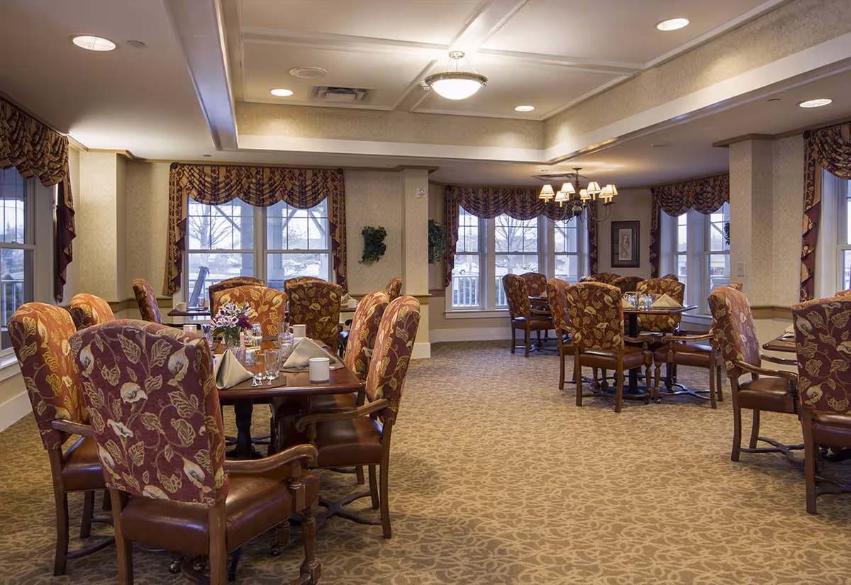 A spacious dining room with multiple wooden tables and cushioned chairs featuring a floral pattern. The tables are set with napkins, glasses, and small flower arrangements. Large windows with patterned valances allow natural light to fill the room. The ceiling has recessed lighting and a chandelier, and the carpet has a subtle floral design.