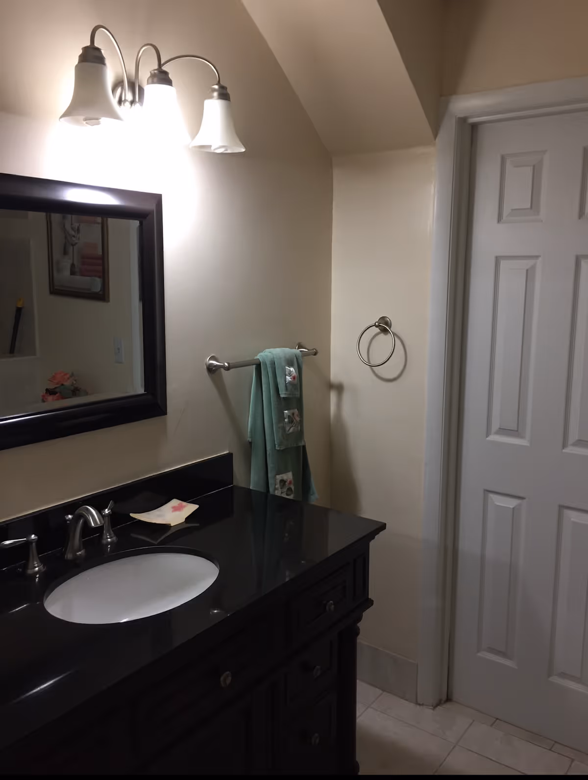 A bathroom interior featuring a black countertop with an oval white sink, a silver faucet, and a dark wooden vanity with drawers. Above the sink is a rectangular mirror with a dark frame and a three-light fixture with frosted glass shades. A towel rack on the wall holds two green towels with decorative patches, and there is an empty towel ring next to a white paneled door.