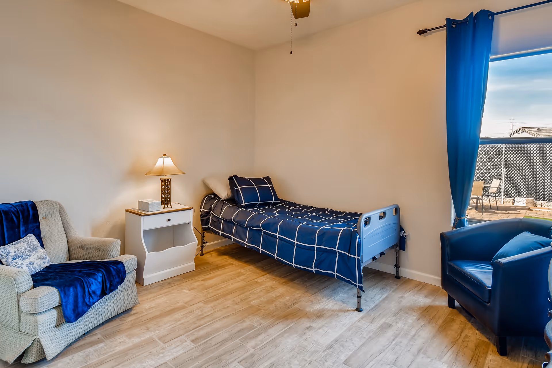 A simple, clean bedroom in an assisted living facility with a single bed covered in a blue checkered blanket, a beige armchair with a blue throw, a white nightstand with a lamp and tissue box, and a blue armchair near a window with blue curtains overlooking an outdoor patio area.