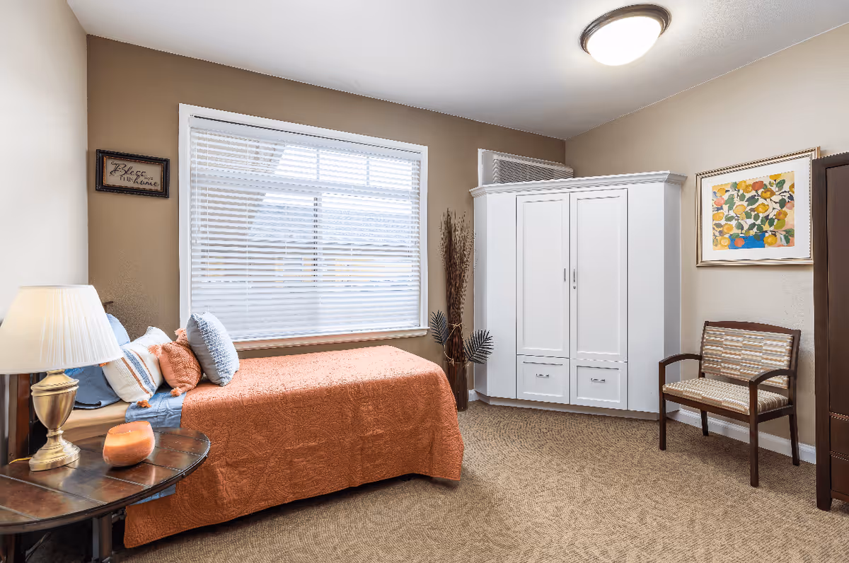 A cozy bedroom with a single bed covered in an orange quilt and multiple pillows. Next to the bed is a wooden side table with a lamp and a small decorative item. The room has a large window with white blinds, a white wardrobe, a wooden chair with a patterned cushion, and a colorful framed painting on the wall. The carpet is beige and the walls are painted in a light tan color.