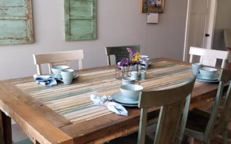 Rustic wooden dining table with mint-green chairs, place settings, napkins and a small floral centerpiece in a bright dining room.