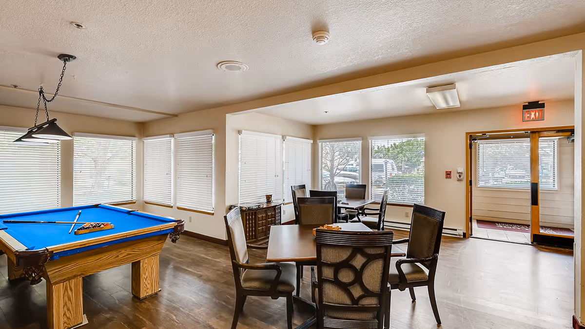 A bright common area with a pool table featuring a blue felt top and a rack of pool balls. There are several tables with chairs arranged around the room. Large windows with blinds allow natural light to fill the space. An exit door with a red exit sign is visible in the background.
