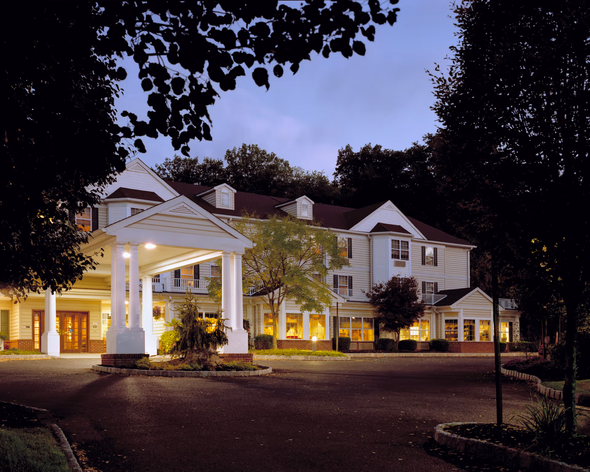 Exterior view of a senior living facility building at dusk with lights glowing from inside. The building has white siding, multiple windows, and a covered entrance supported by white columns. Trees and landscaping surround the driveway in front of the building.