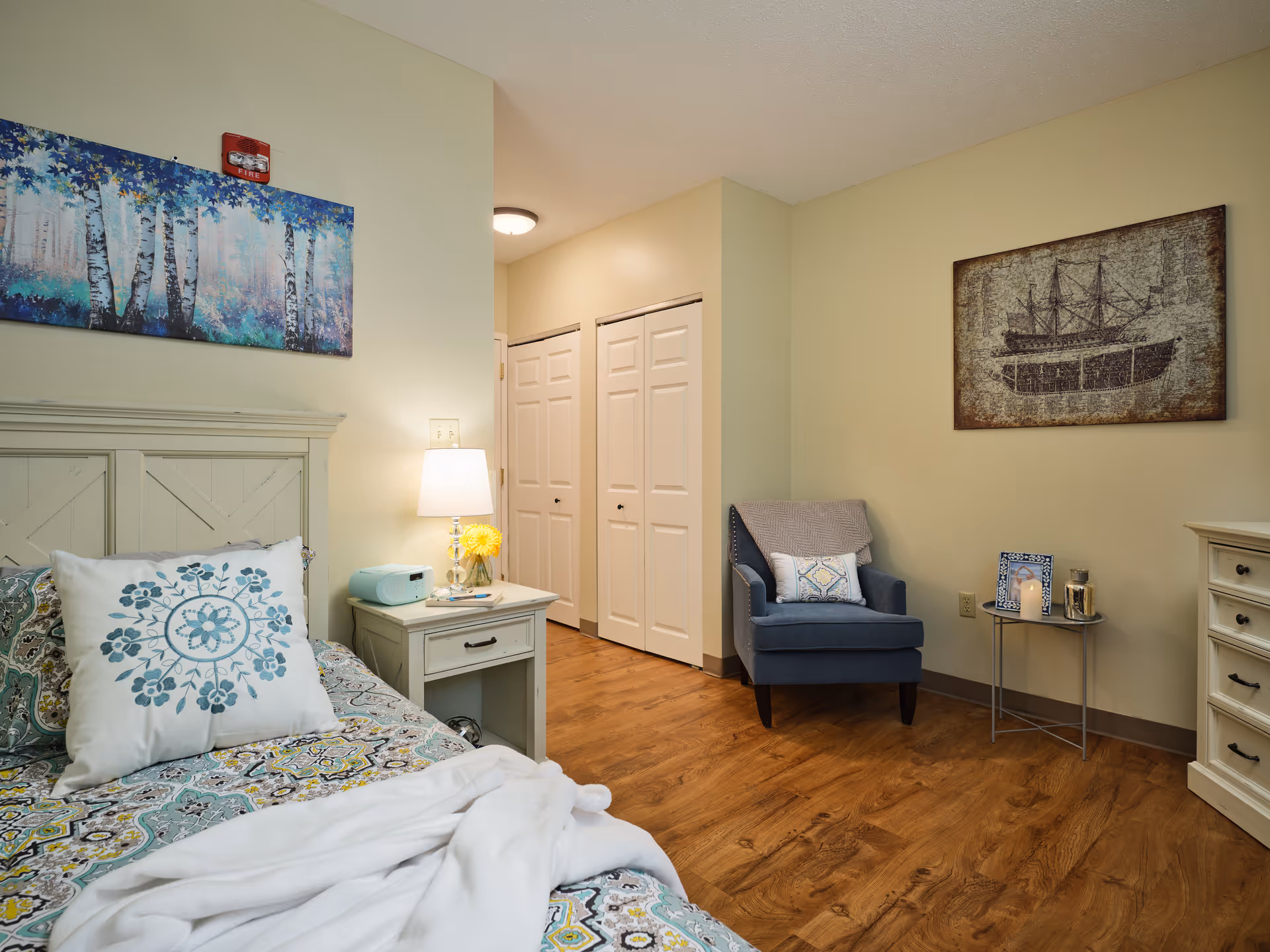 A cozy bedroom with a bed featuring patterned bedding and a decorative pillow. Next to the bed is a white nightstand with a lamp, a small radio, and a yellow flower in a vase. There is a blue armchair with a pillow and a blanket draped over it, positioned near a small round table holding a candle and a framed photo. The room has wooden flooring, light-colored walls, and two pieces of wall art: one depicting birch trees and the other an old ship.