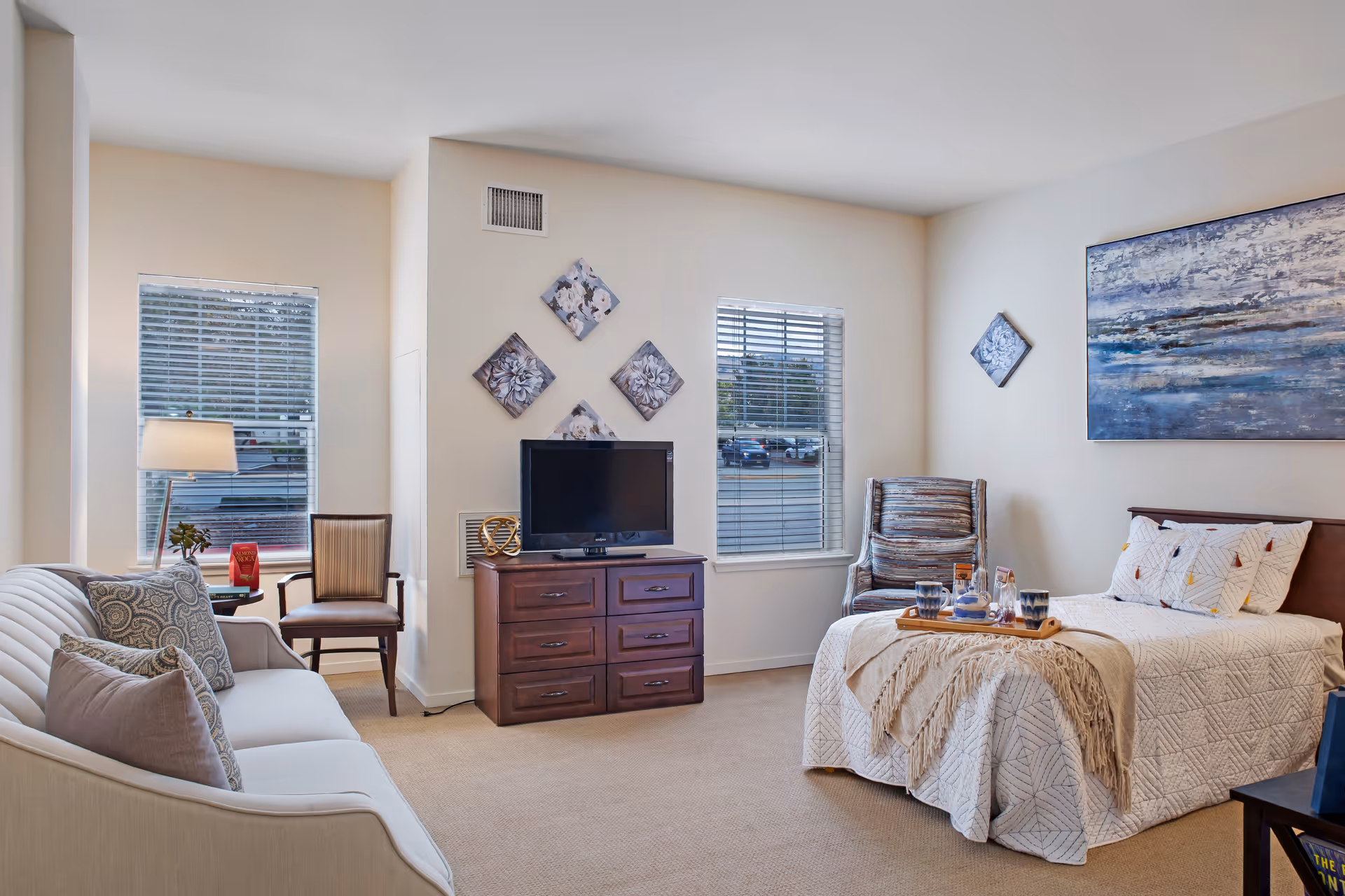 A cozy senior living room featuring a white sofa with patterned pillows, a wooden chair, a TV on a wooden dresser, and a bed with a white quilt and decorative pillows. The room has two windows with blinds, wall art above the TV and bed, and a tray with cups and a teapot on the bed.