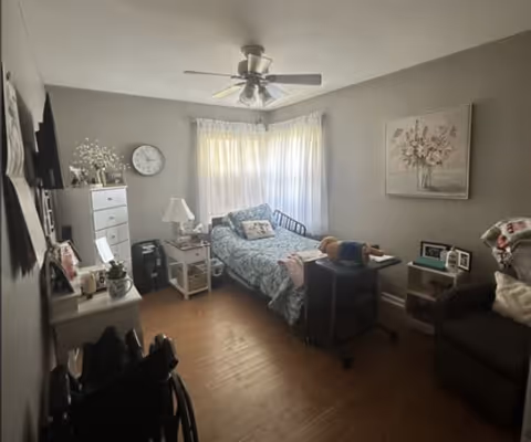 A small, cozy bedroom with a single bed covered in a blue patterned bedspread. There is a bedside table with a lamp and various items, a white dresser with decorative items on top, a clock on the wall, and a ceiling fan overhead. A wheelchair is partially visible in the foreground, and a comfortable armchair with a blanket is placed near the bed. The room has wooden flooring and light gray walls with a floral painting hanging on one wall. Natural light filters in through sheer curtains covering a window.