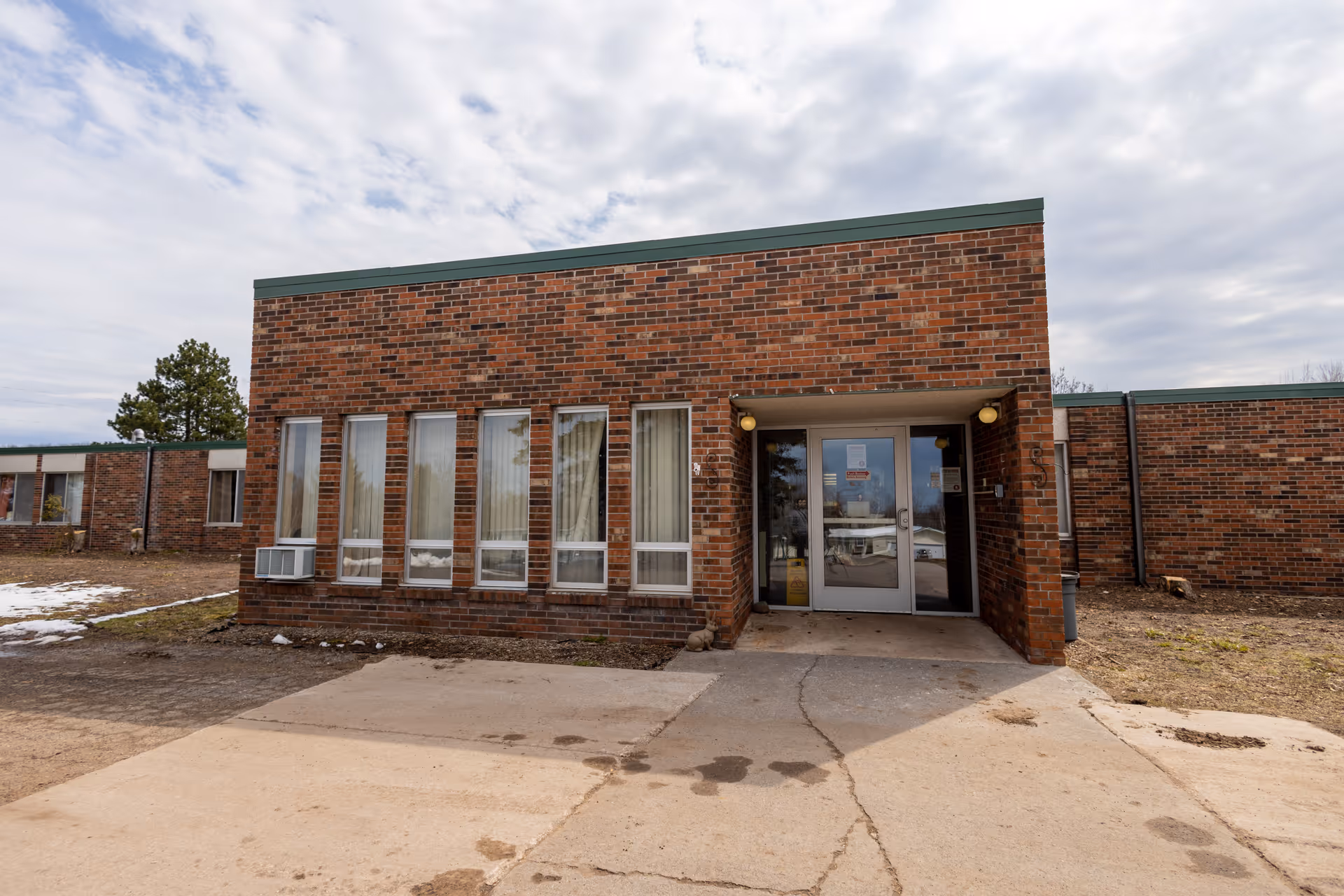 Exterior view of a single-story brick building with multiple vertical windows and a glass door entrance under a small covered area. The ground is paved with concrete and there are patches of dirt and snow around the building. The sky is cloudy.
