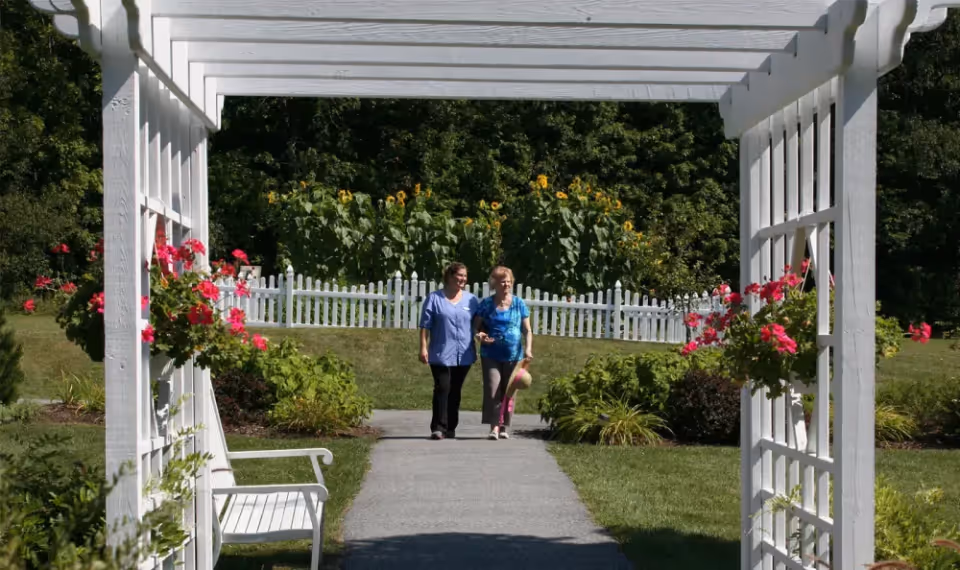 Two women walking arm in arm on a paved path through a garden area with a white wooden pergola overhead, pink flowers hanging on the sides, green bushes, a white picket fence, and tall sunflowers in the background.