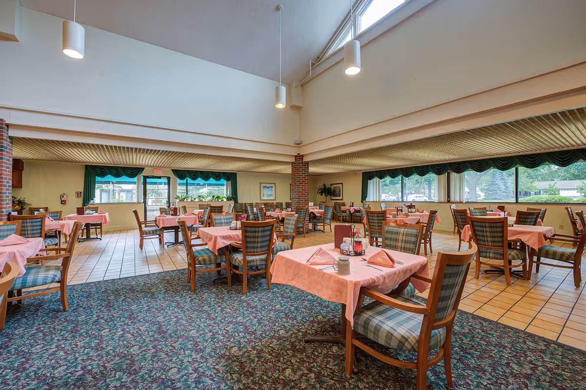 A spacious dining room with multiple tables covered in pink tablecloths and set with napkins, condiments, and utensils. The chairs have wooden frames with plaid upholstery. Large windows with green valances allow natural light to fill the room. The floor is a combination of carpet and tile, and pendant lights hang from the ceiling.