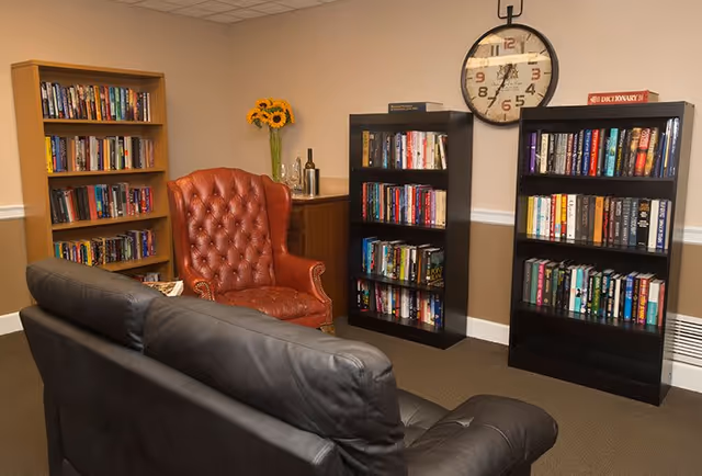 A cozy reading room with a black leather couch facing a red leather armchair. There are three bookshelves filled with books against beige walls. A large round clock hangs on the wall above the bookshelves, and a small table with a vase of sunflowers and a bottle is in the corner.