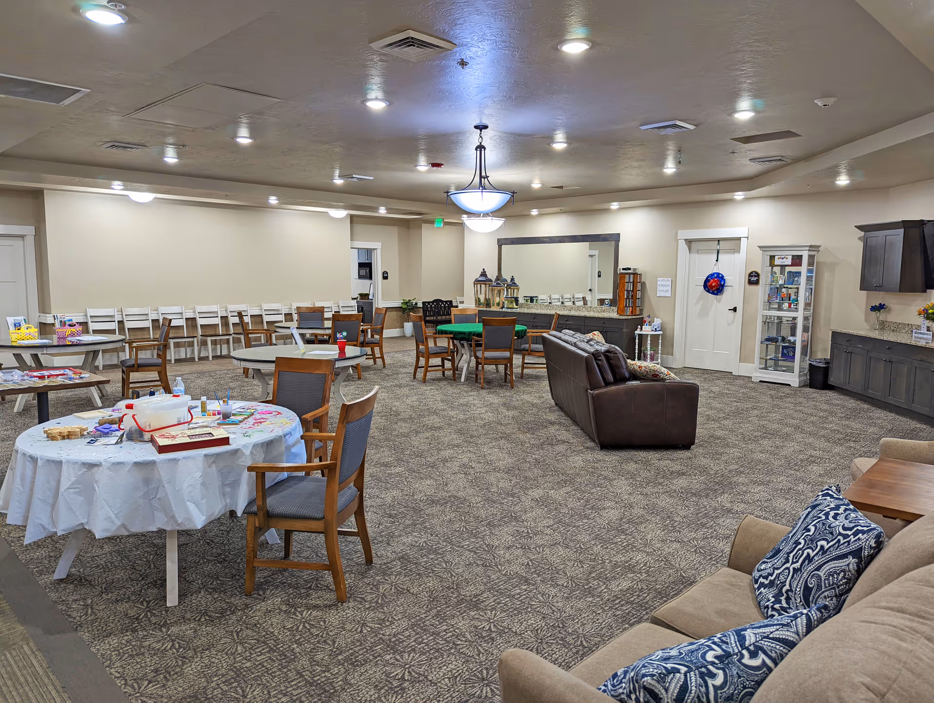 A spacious senior living common area with multiple round tables and chairs, a brown leather couch, and a beige couch with patterned pillows. The room has carpeted floors, neutral-colored walls, ceiling lights, and a large mirror on one wall. There is a cabinet with various items and a door decorated with a wreath.