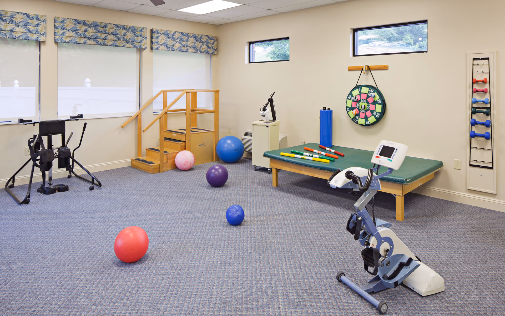 A physical therapy or exercise room with various equipment including an exercise bike, therapy balls, a step platform with handrails, a padded table with colorful rods, a dartboard on the wall, and a rack holding different colored dumbbells. The room has carpeted flooring, beige walls, and windows with patterned valances.