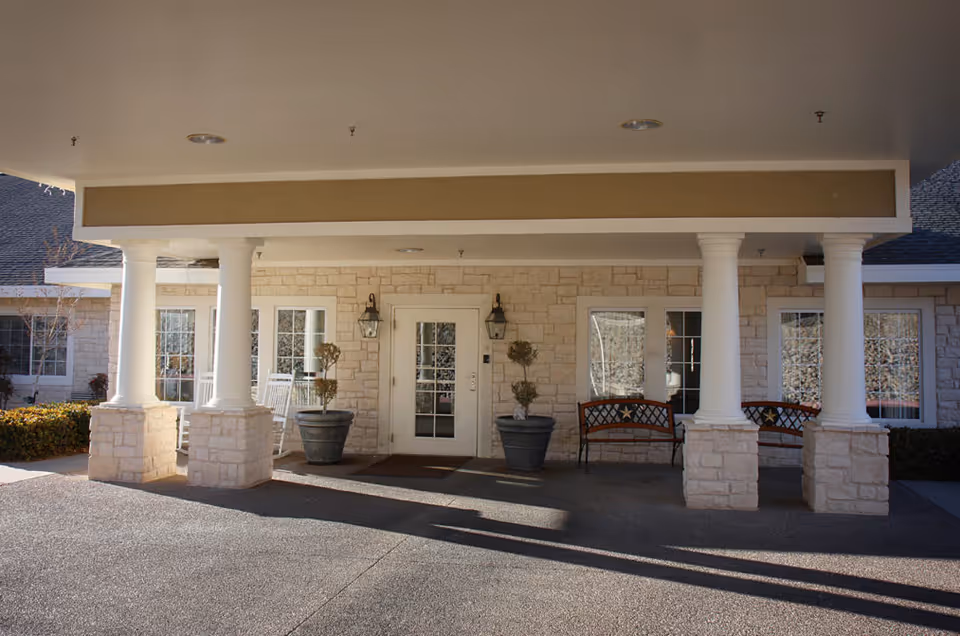 Covered entrance with white columns, double glass doors, potted plants and benches in front of a stone-clad building.
