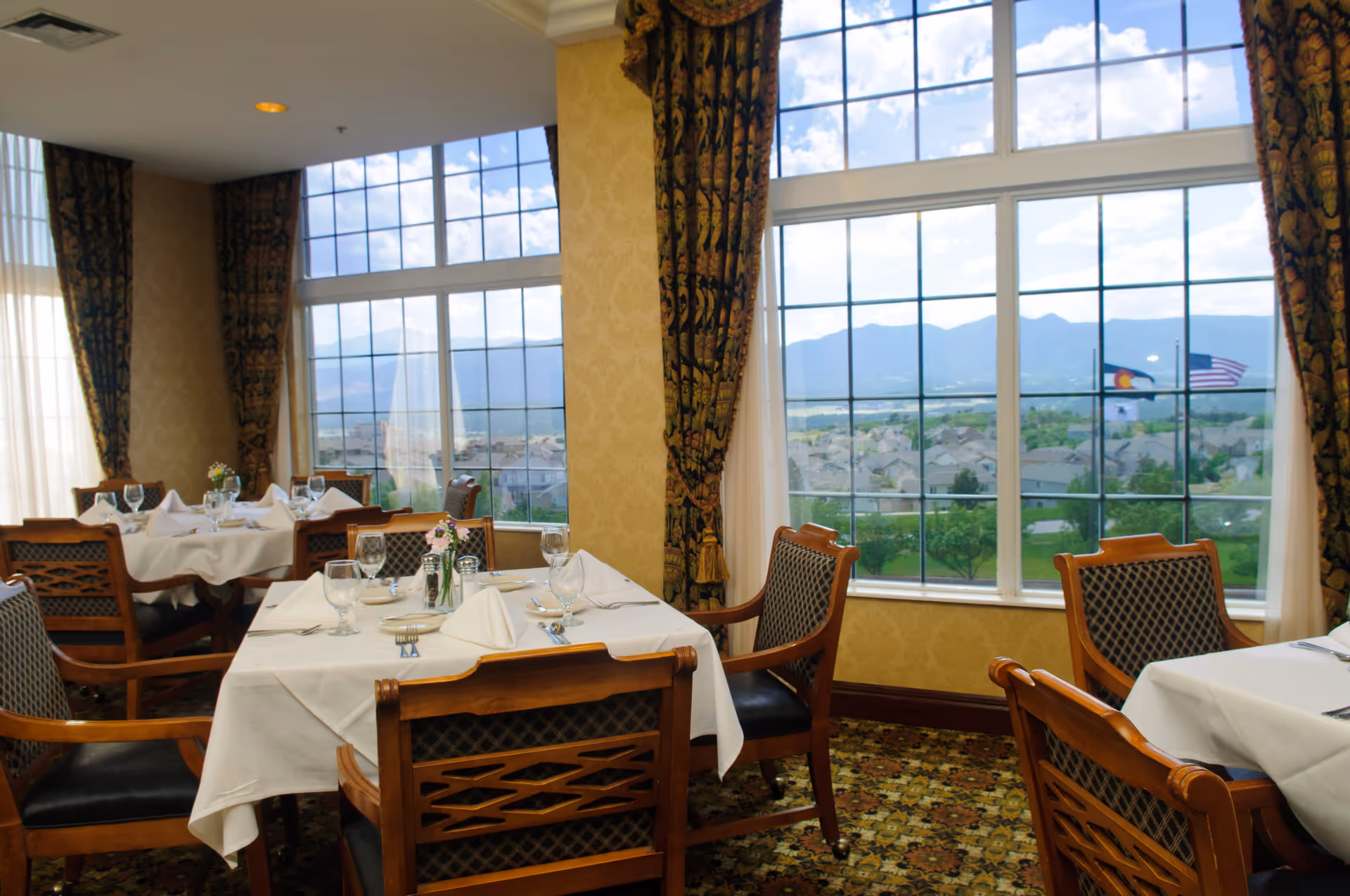A dining room with tables covered in white tablecloths, set with glassware, napkins, and silverware. The room has large windows with a scenic view of a residential area, mountains, and two flags outside. The walls are decorated with patterned wallpaper and heavy curtains.