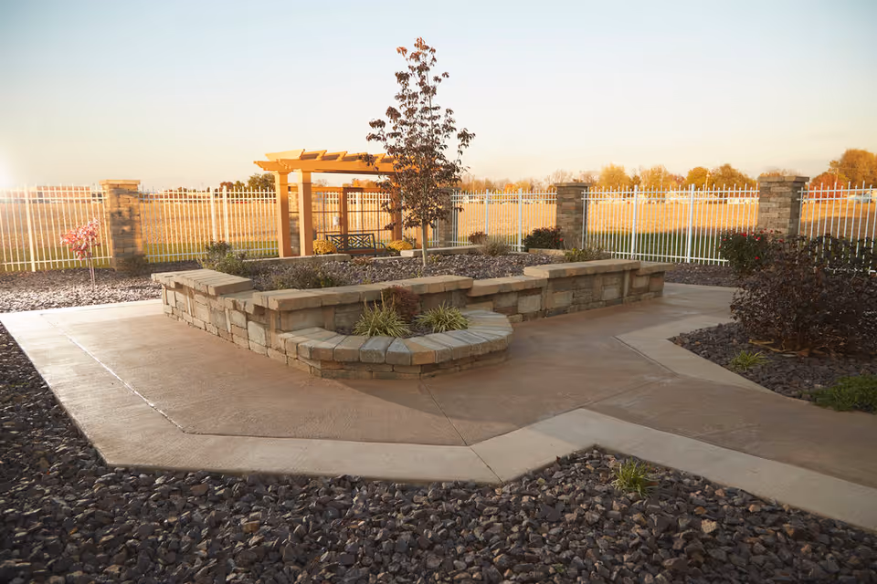 Sunlit outdoor courtyard with stone seating walls, paved walkways, a pergola, and a white fence surrounding landscaped gravel beds.