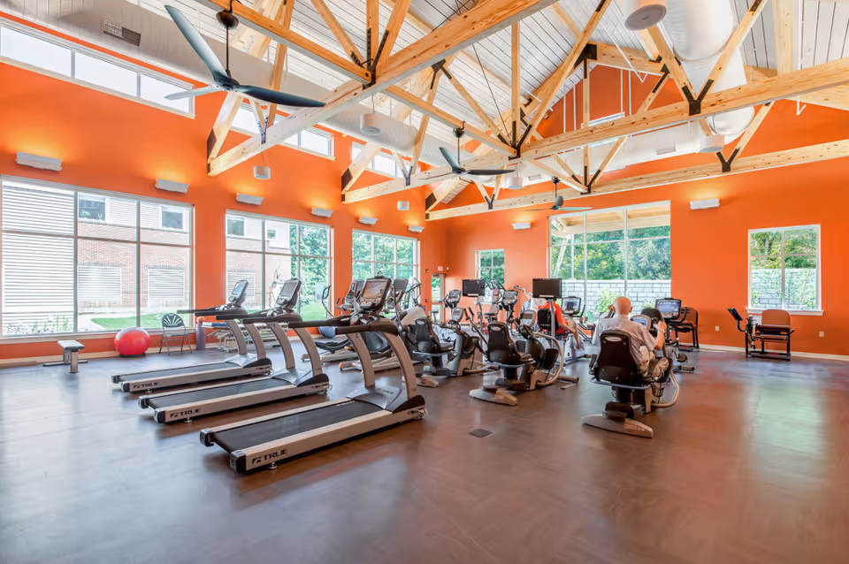A spacious fitness room with large windows letting in natural light, featuring several treadmills and stationary exercise bikes. The room has bright orange walls, exposed wooden beams on the ceiling, and ceiling fans. Two elderly individuals are using the exercise bikes.