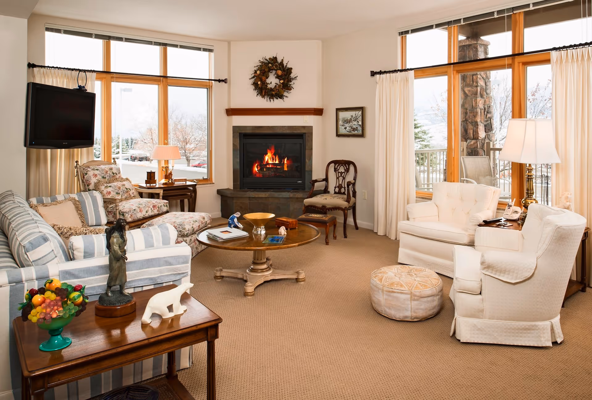 A cozy living room with large windows letting in natural light. The room features a striped sofa, a floral armchair with matching ottoman, two white armchairs, a round wooden coffee table, and a side table with decorative items including a fruit bowl and a small statue. A fireplace with a wreath above it is centered on one wall, and a flat-screen TV is mounted near the windows. Light-colored curtains frame the windows, and a beige carpet covers the floor.