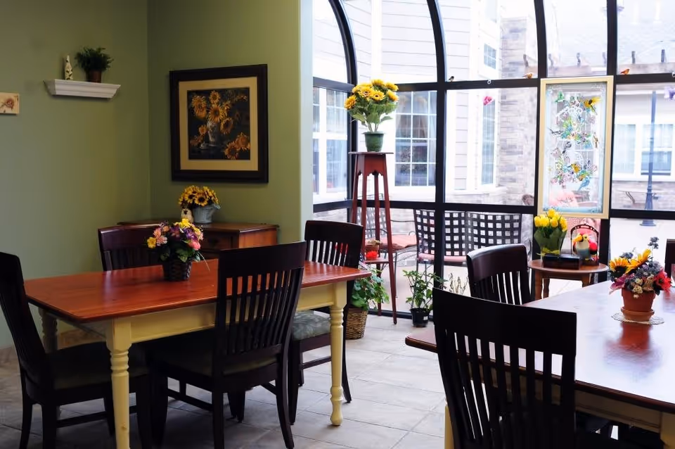 A bright dining area with wooden tables and dark wooden chairs. The tables have flower arrangements in pots. There is a large window with black frames letting in natural light and showing an outdoor patio area with chairs. The walls are light green with a framed sunflower painting and a small white shelf with a plant on it.