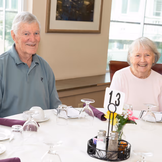 An elderly man and woman sitting at a dining table set with glasses, plates, and napkins, smiling at the camera in a bright room with large windows.