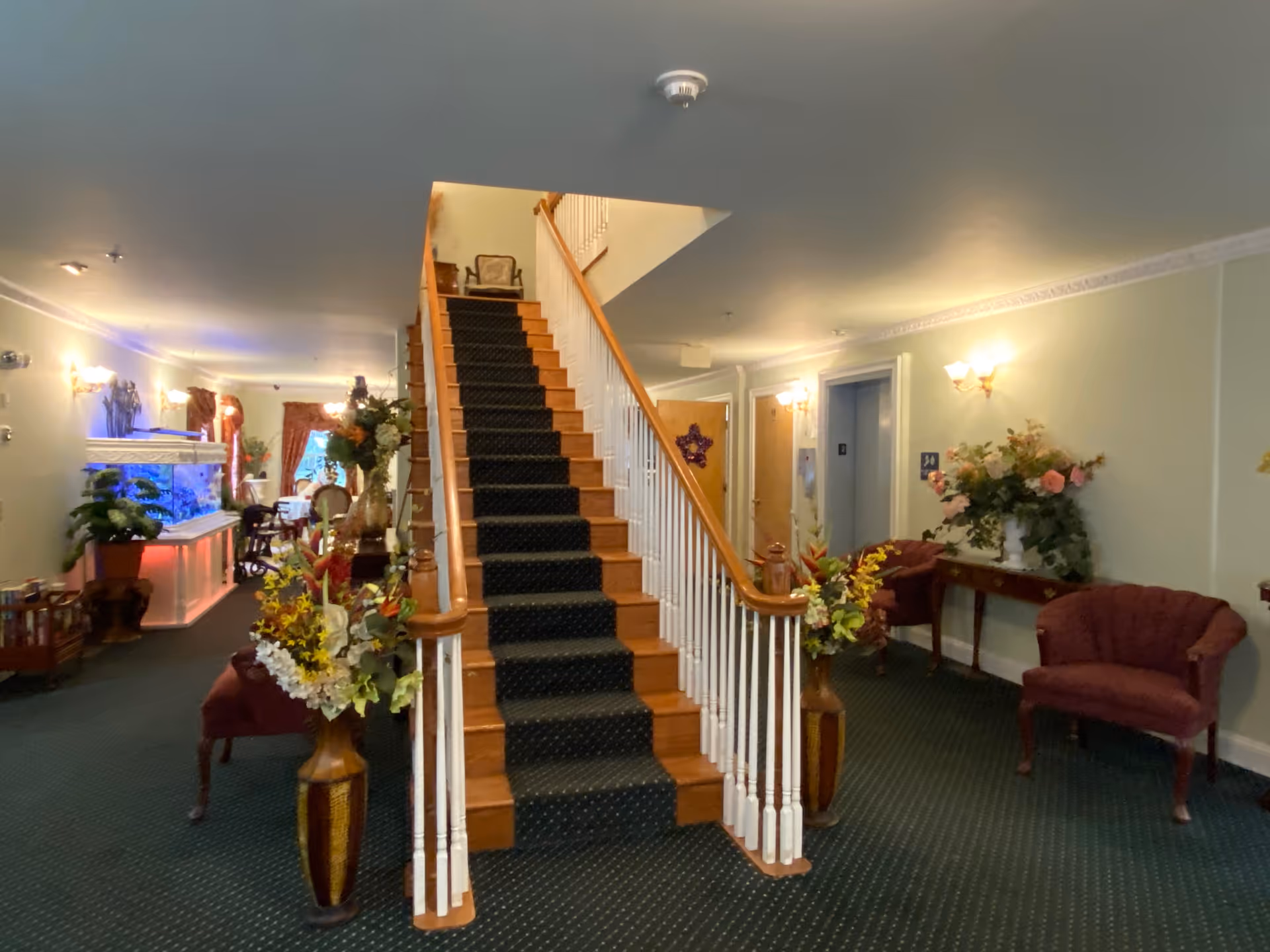Interior view of a senior living facility lobby area featuring a central wooden staircase with a dark carpet runner. The space is decorated with floral arrangements in vases on either side of the staircase and along the walls. There are upholstered chairs and a console table with more flowers. To the left, there is a large fish tank with blue lighting and a seating area with tables and chairs near windows with red curtains. The walls are light-colored with wall sconces providing warm lighting.