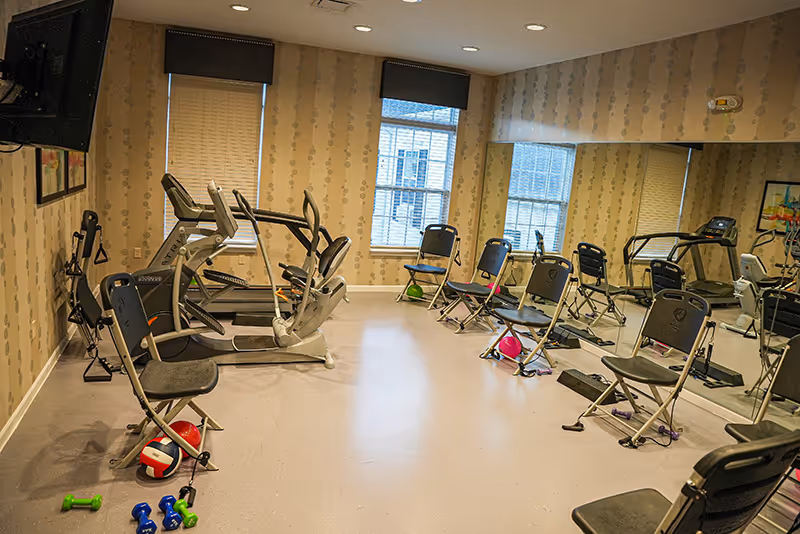 A small fitness room with exercise equipment including treadmills, stationary bikes, and several folding chairs arranged around the room. Various small exercise accessories such as dumbbells, resistance bands, and balls are scattered on the floor. The room has patterned wallpaper, two windows with blinds, and a large mirror covering one wall.
