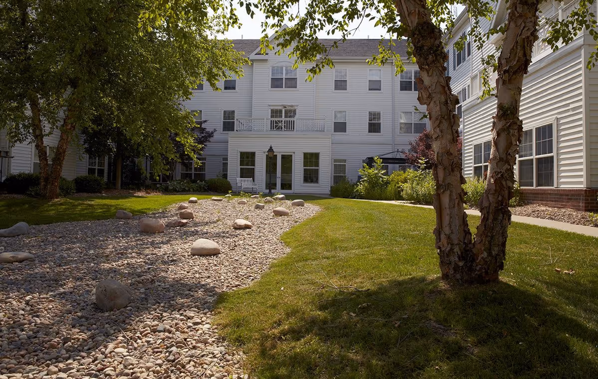 Courtyard of a white multi-story senior living building with a dry rock bed, grassy lawn, trees, and a pathway.