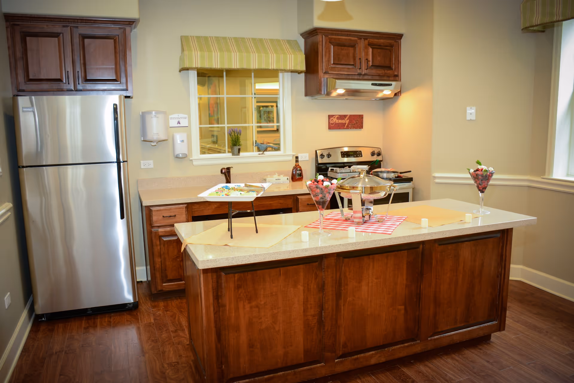 A kitchen area with wooden cabinets and a stainless steel refrigerator on the left. There is a countertop island in the center with a chafing dish, two tall glasses filled with fruit, and a tray with food. The stove and oven are against the back wall under a wooden cabinet and a vent hood. A window with a green striped valance is above the back counter, and the walls are painted a light beige color.