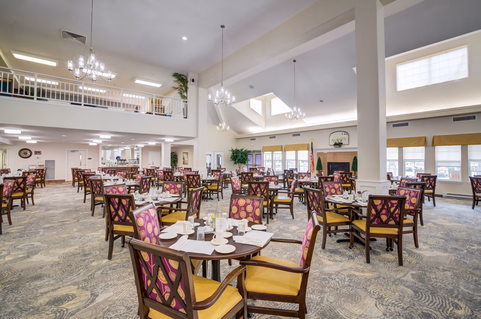 A spacious dining room with multiple round tables set with white plates, cups, and napkins. The chairs have wooden frames with yellow seats and purple patterned backs. The room features high ceilings with chandeliers, large windows with yellow valances, and a fireplace with decorative plants on either side. There is a balcony area above and a carpeted floor with a swirling pattern.