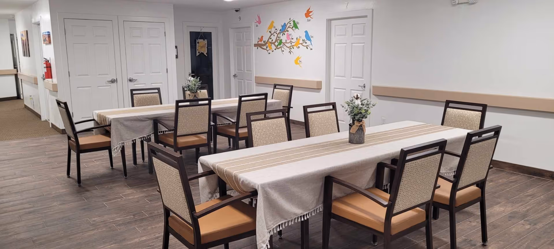 A clean and well-lit dining area with two rectangular tables covered with beige tablecloths and decorated with small flower arrangements. The room has wooden flooring, white walls, several closed white doors, and a colorful bird mural on one wall. There are eight chairs around the tables, and the space appears organized and inviting.